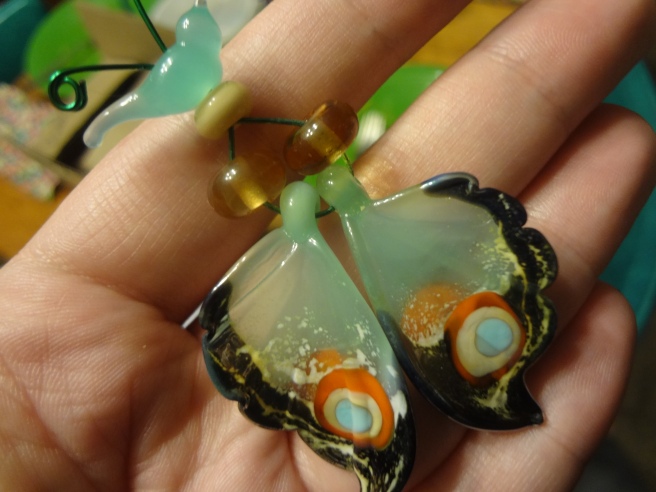 A hand holding a green bird bead, three tan glass beads, and 2 green and brown butterly wings make of glass, all strung on wire.